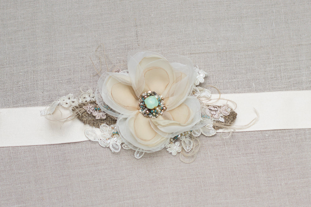A unique handmade wedding dress belt sash features champagne flower, gold, greenish crystals, burlap accents. Floral design bridal sash belt is perfect for rustic, spring, or summer weddings. An online boutique with one-of-a-kind wedding accessories.