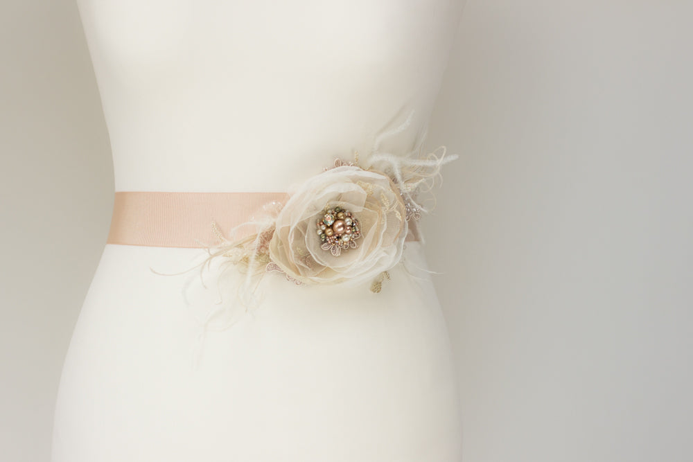  A handmade wedding dress bridal sash belt with a champagne flower, feathers, sage green, and rose gold accents, perfect for rustic, spring, or summer weddings. Find unique wedding accessories at this online boutique. 